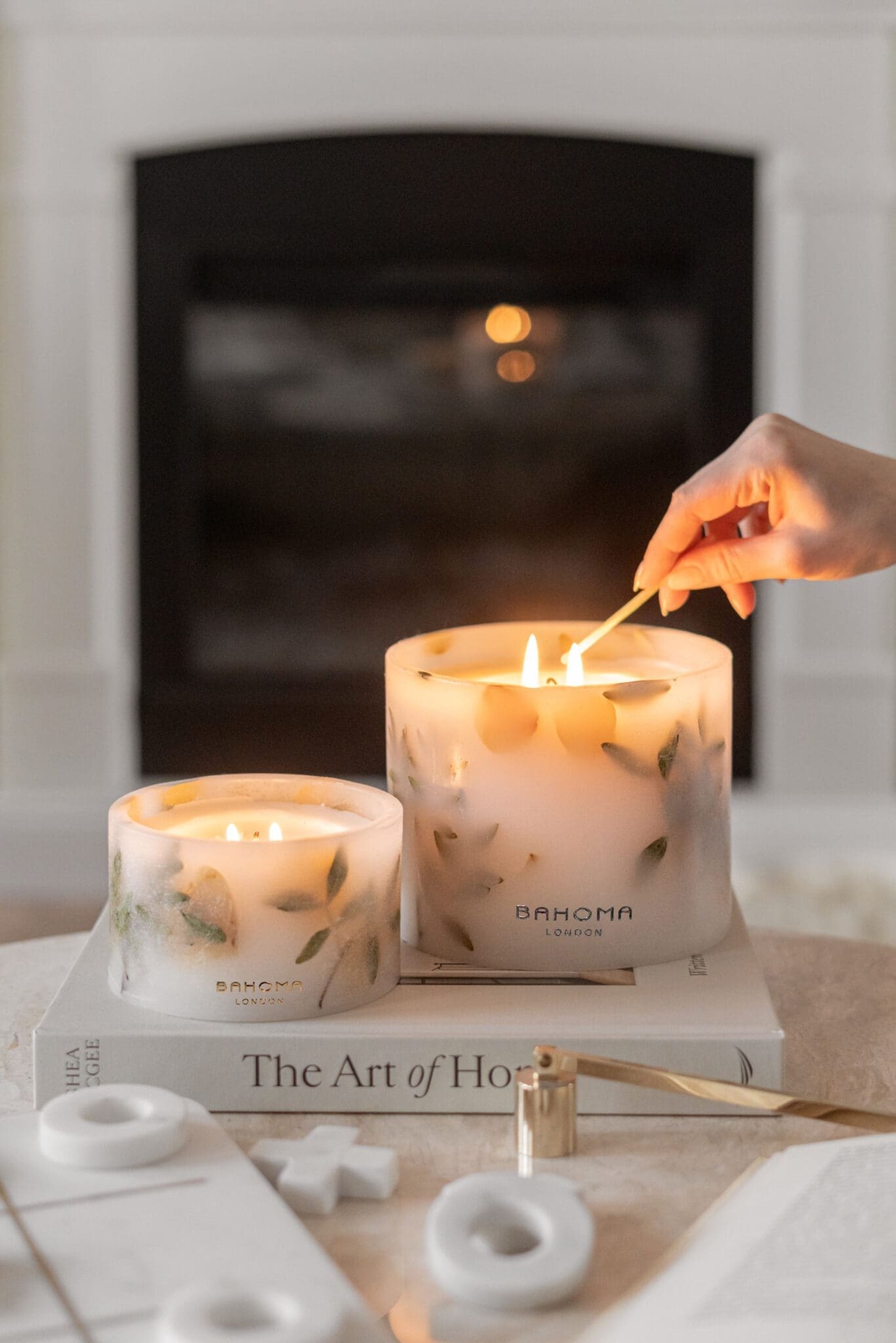 Comparison of Small and Grande Amalfi Coast Scented Pillar Candles side by side. Both displayed on a book on a coffee table in a luxurious, design led living space. Visible hand lighting up the candles.