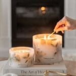 Comparison of Small and Grande Amalfi Coast Scented Pillar Candles side by side. Both displayed on a book on a coffee table in a luxurious, design led living space. Visible hand lighting up the candles.